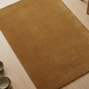 May include: A rectangular, solid-colored rug in a warm, golden-brown hue. The rug has a soft, plush texture and is placed on a light-colored wooden floor. The rug's simple design and neutral color make it suitable for various interior styles.