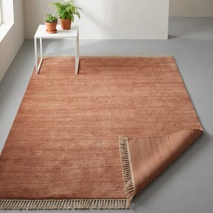 May include: A terracotta-coloured rectangular rug with a textured surface and fringed edges. The corner of the rug is folded up, revealing the underside. A small white table with potted plants is in the background.