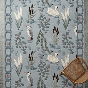 May include: A pale blue rug depicting a pond scene with swans, herons, and reeds. The rug has a chain-link border with pink floral details. A woven chair is in the corner.