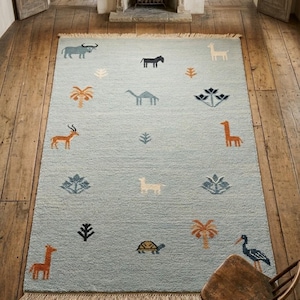 May include: A light blue rectangular rug with a fringed edge. The rug features various animal and plant designs in shades of brown, black, and blue. The animals include a giraffe, camel, and turtle.