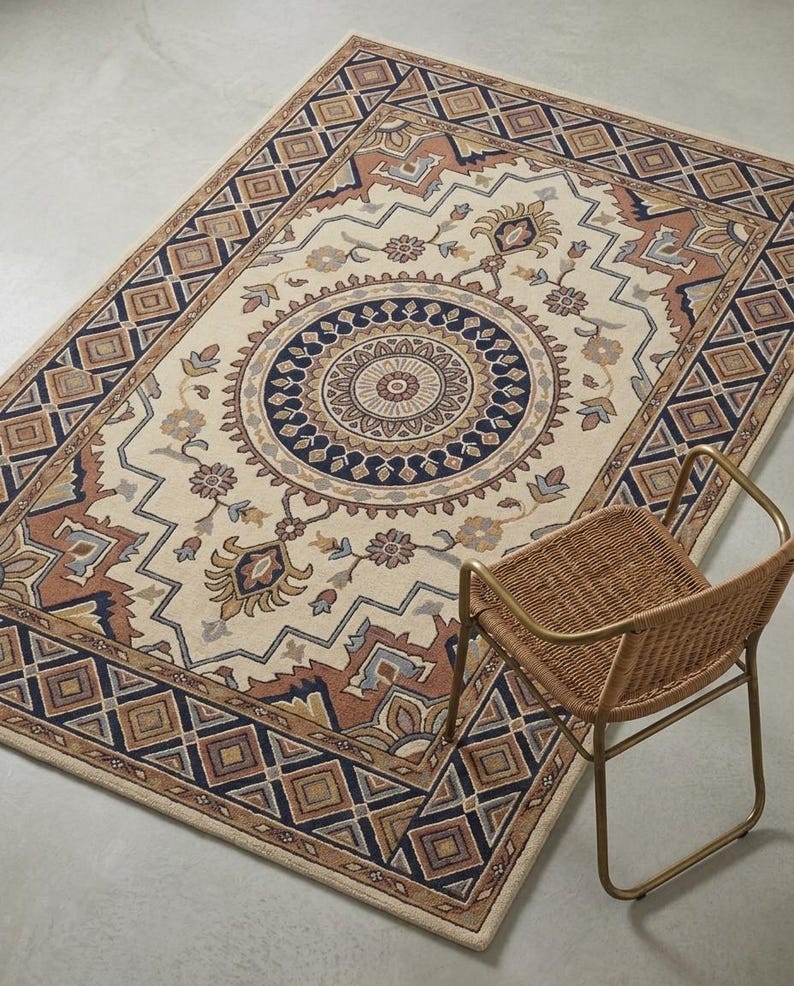 May include: Rectangular beige rug with a central medallion design in navy blue, brown, and tan. The rug features a geometric border and floral accents. A wicker chair with a gold metal frame sits beside the rug.