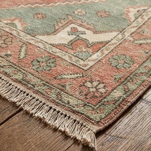 May include: Close-up of a distressed, rectangular rug with a traditional pattern. The rug features a salmon-colored field with a central section in muted green. The design includes floral and geometric motifs, with a fringed edge.
