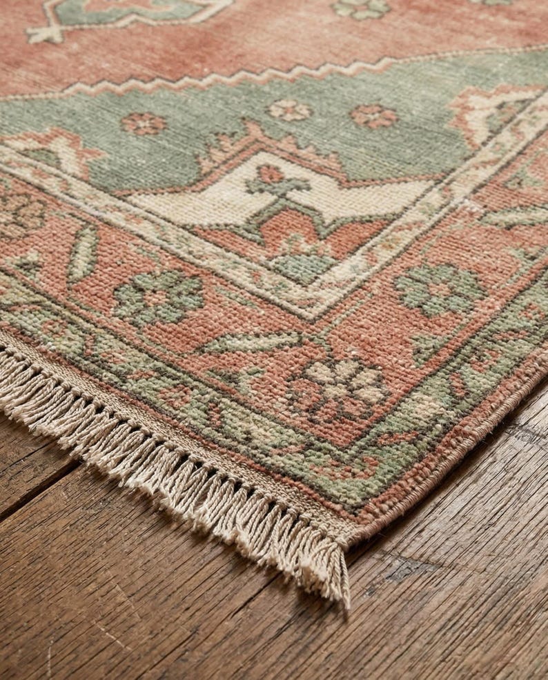 May include: Close-up of a distressed, rectangular rug with a traditional pattern. The rug features a salmon-colored field with a central section in muted green. The design includes floral and geometric motifs, with a fringed edge.