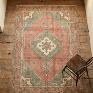 May include: A rectangular, distressed-look rug with a central diamond pattern. The rug features a color palette of muted coral, sage green, and cream. The rug is placed on a wooden floor, with a wooden chair in the corner.
