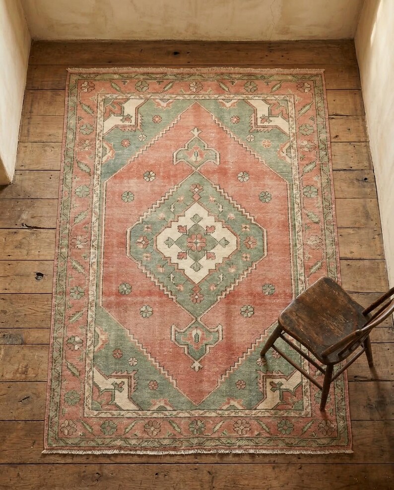 May include: A rectangular, distressed-look rug with a central diamond pattern. The rug features a color palette of muted coral, sage green, and cream. The rug is placed on a wooden floor, with a wooden chair in the corner.