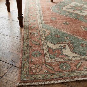 May include: Close-up of a distressed, rectangular rug with a traditional pattern. The rug features a faded red-orange field with geometric designs in muted green and cream. The border has floral motifs. The rug is placed on a wooden floor.
