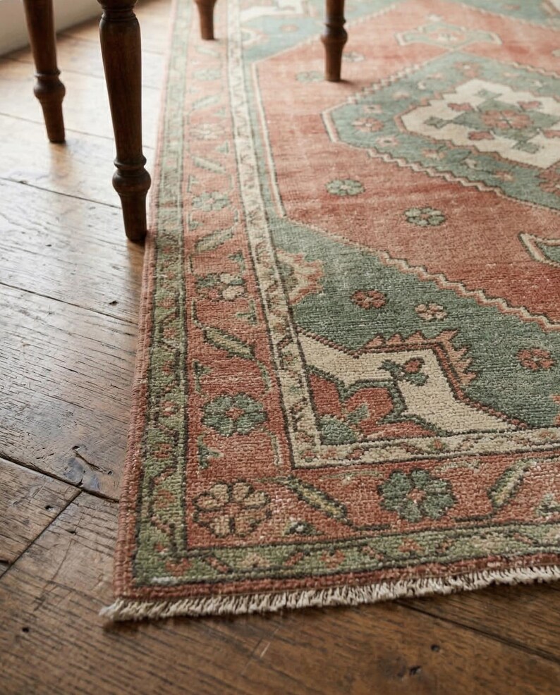 May include: Close-up of a distressed, rectangular rug with a traditional pattern. The rug features a faded red-orange field with geometric designs in muted green and cream. The border has floral motifs. The rug is placed on a wooden floor.