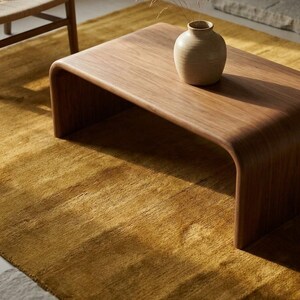 May include: A contemporary, curved wooden coffee table in a light brown hue. A small, beige ceramic vase with dried grass is placed on the table. The table rests on a golden-yellow rug, with a woven chair in the background.