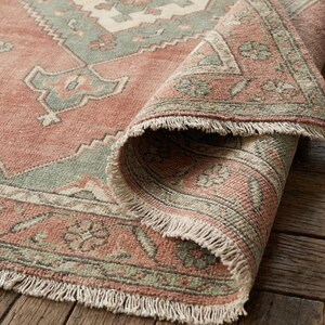 May include: A rolled-up, distressed-look rug with a faded red background and a geometric pattern in shades of green and cream. The rug has a fringed edge and is displayed on a wooden surface.