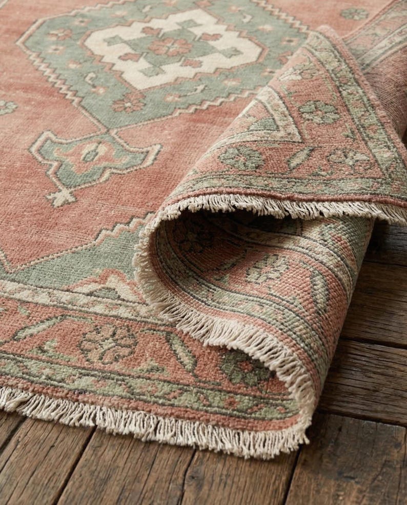 May include: A rolled-up, distressed-look rug with a faded red background and a geometric pattern in shades of green and cream. The rug has a fringed edge and is displayed on a wooden surface.