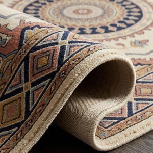May include: Rolled-up area rug featuring a cream backdrop and an intricate geometric design. The pattern includes navy blue, peach, and gold hues. The rug's edge shows a beige backing and a fringed border, showcasing the rug's construction.