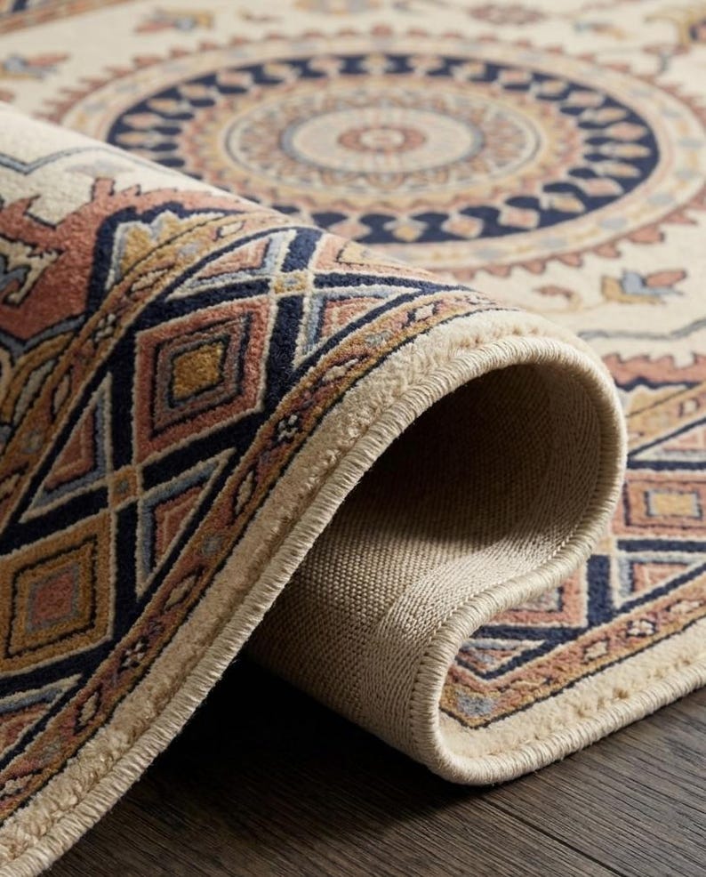 May include: Rolled-up area rug featuring a cream backdrop and an intricate geometric design. The pattern includes navy blue, peach, and gold hues. The rug's edge shows a beige backing and a fringed border, showcasing the rug's construction.