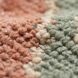 May include: Close-up of a textured rug with a repeating pattern. The rug features a mix of colors, including shades of peach, sage green, and cream. The fibers are tightly woven, creating a soft, plush appearance. The overall design is abstract and modern.
