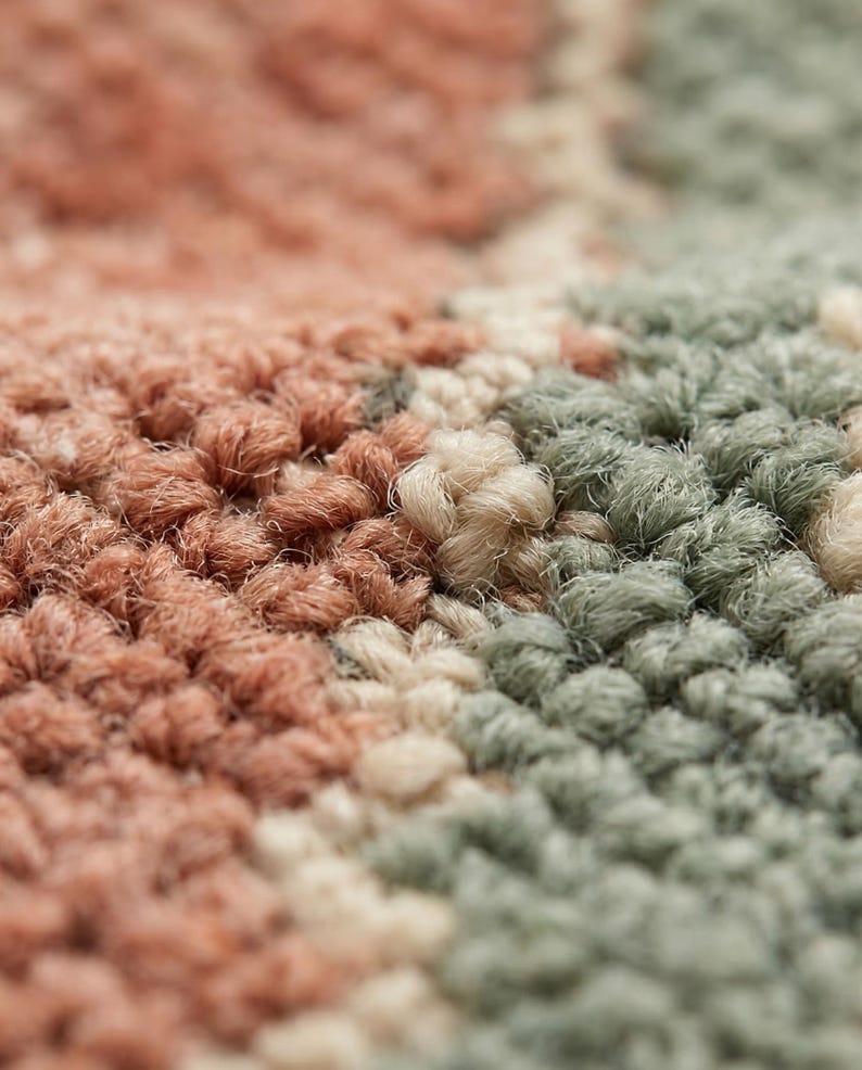 May include: Close-up of a textured rug with a repeating pattern. The rug features a mix of colors, including shades of peach, sage green, and cream. The fibers are tightly woven, creating a soft, plush appearance. The overall design is abstract and modern.