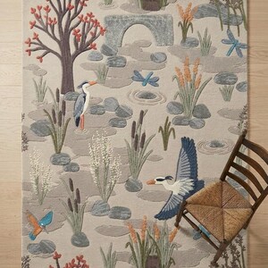 May include: A rectangular rug depicting a pond scene with birds, reeds, and a stone bridge. The colour scheme includes beige, grey, brown, orange, and blue. A wooden chair is positioned on the right side of the image.