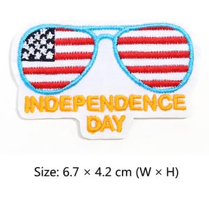 Puede incluir: Parche bordado con gafas de sol con el diseño de la bandera estadounidense. Las gafas de sol están delineadas en azul, y el parche incluye el texto "INDEPENDENCE DAY" en amarillo. El parche mide 6,7 x 4,2 cm.
