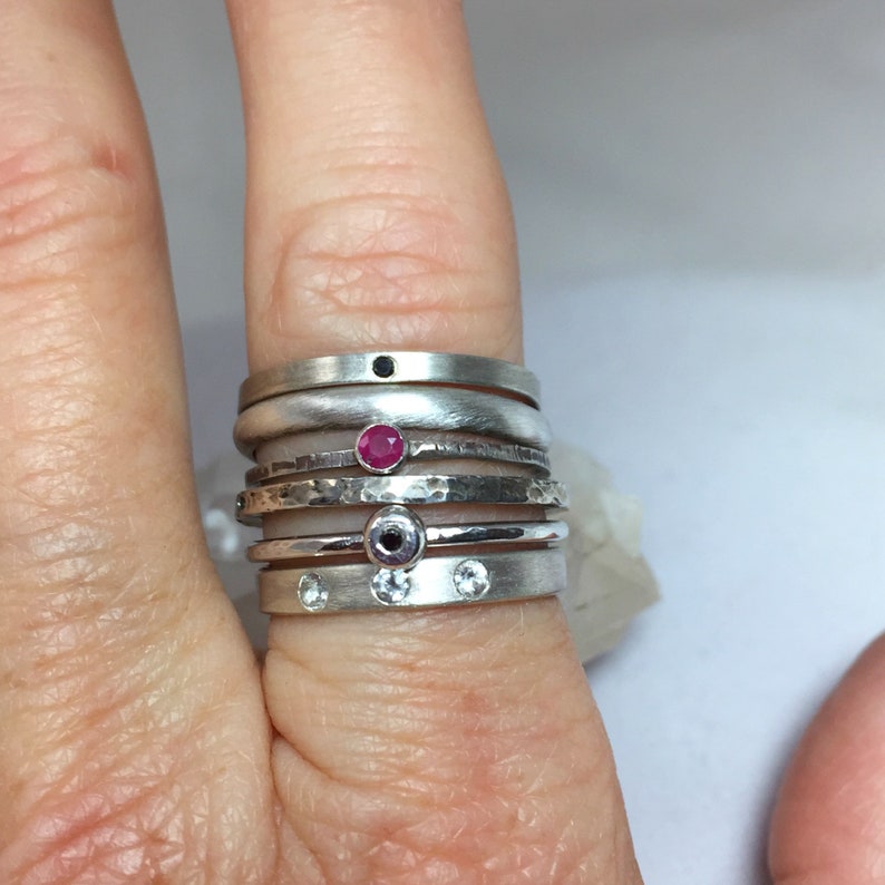 May include: A stack of silver rings with different designs, including a hammered band, a plain band, and a band with a small black stone. The top ring has a small red stone.