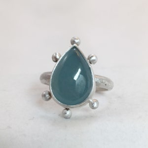 Op de afbeelding: Een zilveren ring met een traanvormige blauwe edelsteen gezet in een zilveren bezel. De ring heeft zes kleine zilveren balletjes rondom de edelsteen.