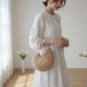 May include: A beige, crocheted handbag with a circular design. The bag has a woven handle and a long, pearl-like strap. The bag is held by a person wearing a white dress.