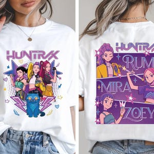 Puede incluir: Camiseta blanca con un gráfico colorido de un grupo de mujeres animadas y el texto "HUNTRAX". El diseño frontal incluye un gato azul y estrellas. La parte posterior de la camiseta muestra el texto "HUNTRAX" y los nombres "RUMI", "MIRA" y "ZOEY".