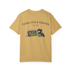 May include: Mustard yellow t-shirt with a graphic featuring a dachshund dog and a record player. The text "TECKEL FUR & FEATHER EST. 2025" is printed above the graphic. The dog is black and white, and the record player is green and red.