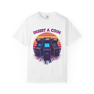Op de afbeelding: Wit T-shirt met een retro arcade game design. De afbeelding toont drie arcadekasten tegen een zonsondergang, met de tekst "INSERT A COIN" erboven. Het ontwerp maakt gebruik van een levendig, kleurrijk palet, dat een vintage gevoel oproept.