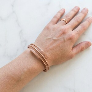Op de afbeelding: Een koperkleurige armband met een textuurontwerp en een bijpassende ring. De armband wikkelt zich om de pols en toont een minimalistische en elegante stijl. De sieraden worden gepresenteerd op een lichtgekleurde achtergrond.