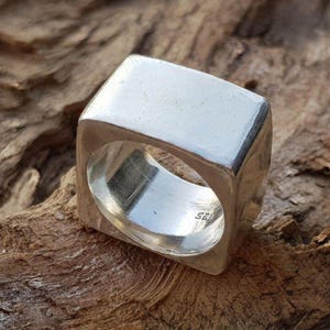 Puede incluir: Un anillo cuadrado de plata con un acabado pulido. El anillo tiene una parte superior rectangular sólida y un interior redondeado y abierto. El anillo está sobre un fondo marrón texturizado.