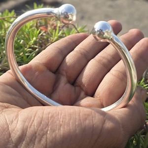 Könnte beinhalten: Ein silberfarbenes Armreif-Armband mit glattem, geschwungenem Design. Das Armband hat an jedem Ende eine abgerundete Kugel. Der Schmuck wird in einer Hand gehalten, mit grünem Laub im Hintergrund.