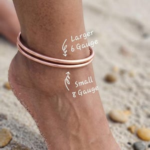 Puede incluir: Tobillera de oro rosa con dos bandas, etiquetada como "Larger 6 Gauge" y "Small 8 Gauge". La tobillera se lleva en un tobillo en una playa de arena. La imagen muestra el pie y el tobillo con la joya.