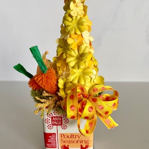 Può includere: Composizione floreale decorativa in una lattina riutilizzata di Ann Page Poultry Seasoning. La composizione presenta un cono di fiori di carta gialli, pompon arancioni e un nastro giallo a pois. La latta è bianca con dettagli rossi.