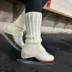 May include: A pair of off-white crocheted leg warmers featuring a vertical ribbed pattern on the upper section and a textured, open-stitch design at the base, worn over white ice skates with silver metal blades and black leggings.