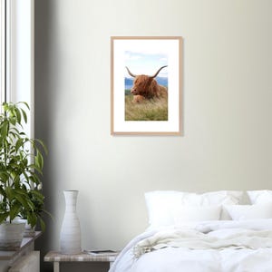 Può includere: Stampa incorniciata raffigurante una mucca delle Highlands, con lunghe corna e pelo marrone-rossiccio, sdraiata nell'erba alta. Lo sfondo mostra un cielo blu e montagne. La cornice è in legno chiaro, ideale per un soggiorno o una camera da letto.