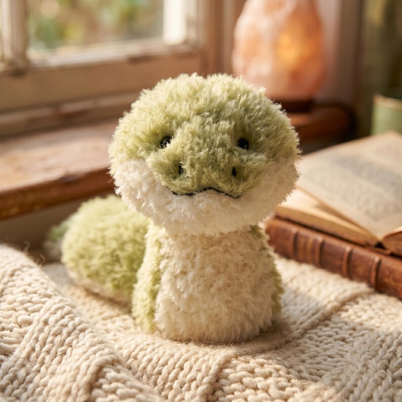 May include: A plush, light green snake toy with a white belly and head. The snake is sitting on a cream-colored knitted blanket. The toy has small black eyes and a stitched smile. The background includes a window and a stack of books.