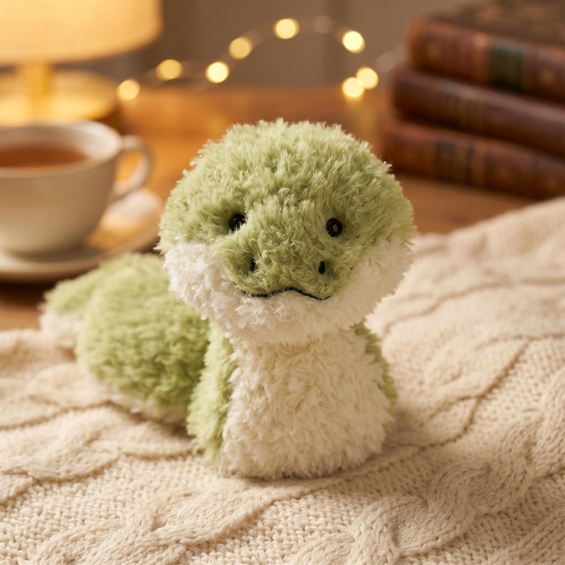 May include: A plush green and white stuffed animal shaped like a reptile, with black eyes and a smiling mouth. The toy is on a cream-colored cable knit blanket. A cup of tea and books are in the background.