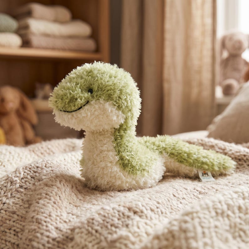 May include: A plush green and white snake toy with a smiling face rests on a beige knitted blanket. The snake has a soft, fuzzy texture and a curved tail. The background includes a wooden shelf with folded towels and a teddy bear.