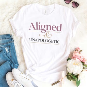 May include: White t-shirt with the words "Aligned & Unapologetic" in mauve and gold lettering. The shirt is styled with blue jeans, white sneakers, and sunglasses. A bouquet of pink and white flowers is also in the image.