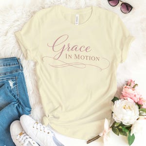 May include: Cream-colored t-shirt with the words "Grace in Motion" in a mauve script. The shirt is styled with blue jeans, white sneakers, and sunglasses. A bouquet of pink and white flowers is also in the image.