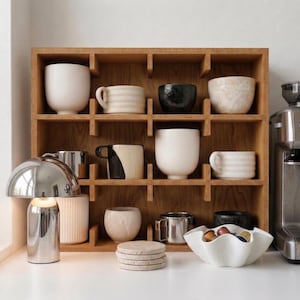 May include: A wooden shelf unit displays various ceramic mugs, cups, and bowls in neutral tones. A silver lamp and a coffee machine are on the counter. Coasters and a white bowl with coffee pods are also visible.