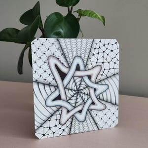 May include: Square artwork with a geometric design in black and white, featuring a central star-like shape in light blue and grey. The artwork has a spiral pattern and is displayed on a pink surface with a green plant in the background.