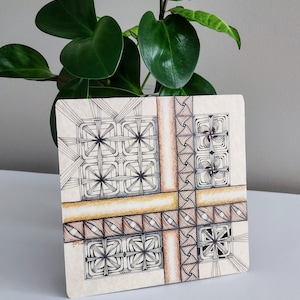 May include: Square artwork with a geometric black and white design, featuring intersecting lines and patterns. The artwork has a beige background with brown and gold accents. A green plant is in the background.