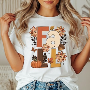 Puede incluir: Una camiseta blanca de manga corta con un estampado gráfico de la palabra Fall en letras grandes. El diseño incluye tonos naranja, marrón y crema con ilustraciones de flores, una calabaza, una bellota, hojas y una mariposa.