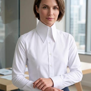 Può includere: Una camicia bianca abbottonata con un colletto alto e strutturato. La camicia a maniche lunghe ha polsini con bottoni e una vestibilità su misura. Il colletto presenta una fila di bottoni. La camicia è indossata con pantaloni blu scuro.