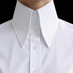 Può includere: Primo piano di una camicia bianca con colletto alto e appuntito. Il colletto è abbottonato con quattro bottoni e la camicia ha un'abbottonatura classica. Il tessuto appare nitido e pulito, con una trama liscia.