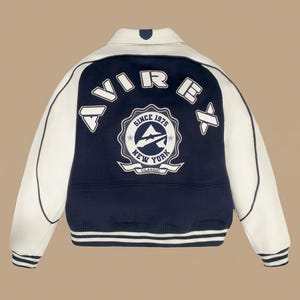 May include: A navy blue and white varsity jacket with the text "AVIREX" across the back. The jacket features a circular emblem with the text "SINCE 1975 NEW YORK CLASSIC". The jacket has white sleeves and a striped hem.