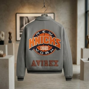 May include: A grey leather varsity jacket featuring a large embroidered New York Knicks logo on the back with orange and navy blue lettering. The word Avirex is embroidered in orange below the logo. The jacket has ribbed cuffs and a collar.