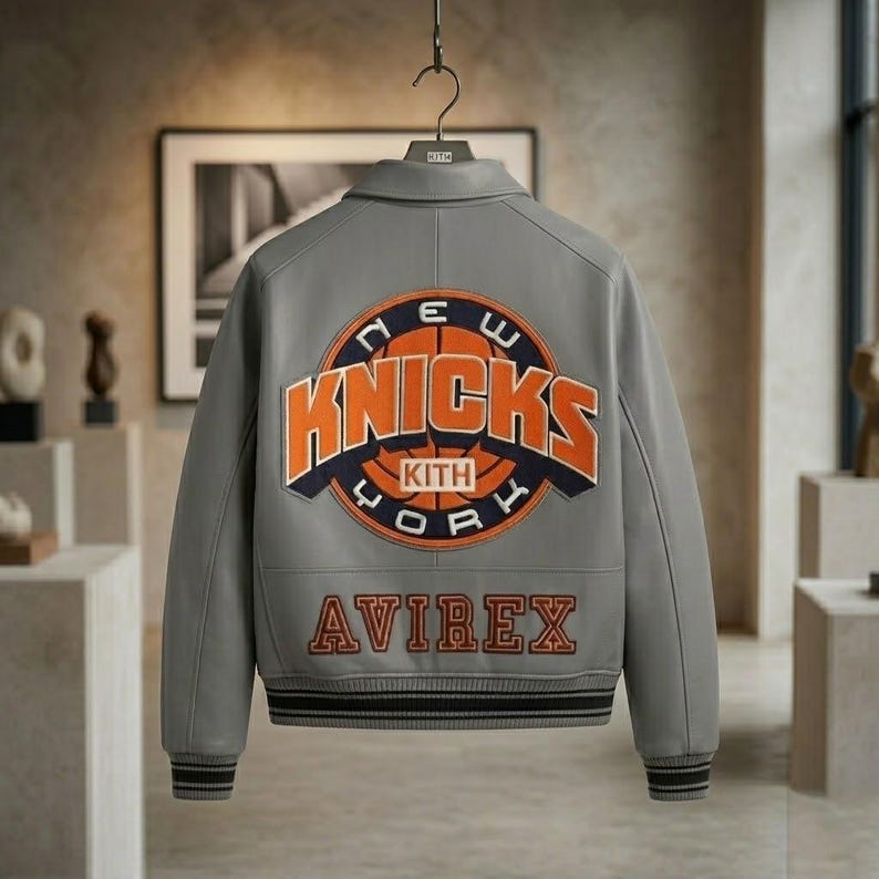 May include: A grey leather varsity jacket featuring a large embroidered New York Knicks logo on the back with orange and navy blue lettering. The word Avirex is embroidered in orange below the logo. The jacket has ribbed cuffs and a collar.