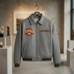 May include: A grey leather jacket featuring a full-length front zipper, a classic collar, and ribbed cuffs and hem. The chest displays an embroidered orange and black New York Knicks basketball logo on the right and the text AVIREX on the left.