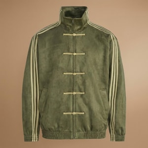 May include: An olive green track jacket with a high collar and a zip. The jacket has three cream-coloured stripes down each sleeve and cream-coloured toggle fastenings. The jacket has a soft, velvety texture. The jacket is a stylish piece of outerwear.