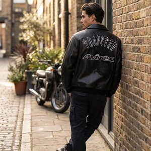 K&ouml;nnte beinhalten: Schwarze Lederjacke mit dem Text "TRAPSTAR" und "Avirex" in Wei&szlig;. Die Jacke hat einen klassischen Bomberstil mit geripptem Kragen und B&uuml;ndchen. Die Person lehnt an einer Backsteinmauer neben einem Motorrad.